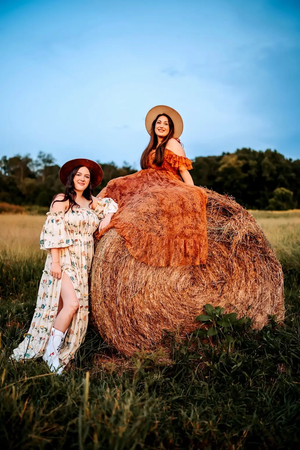 Ashley sitting on top of a round hay bale with her daughter Kayla standing beside it, both in dresses and hats at golden hour